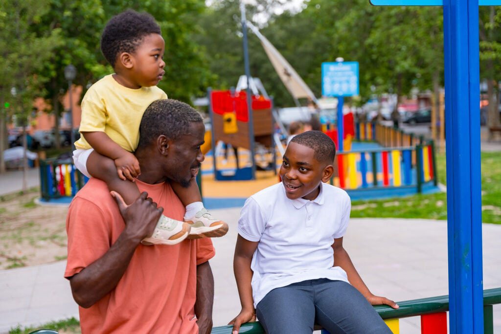 Black father holding toddler on shoulders and smiling at adolescent son sitting on playground equipment