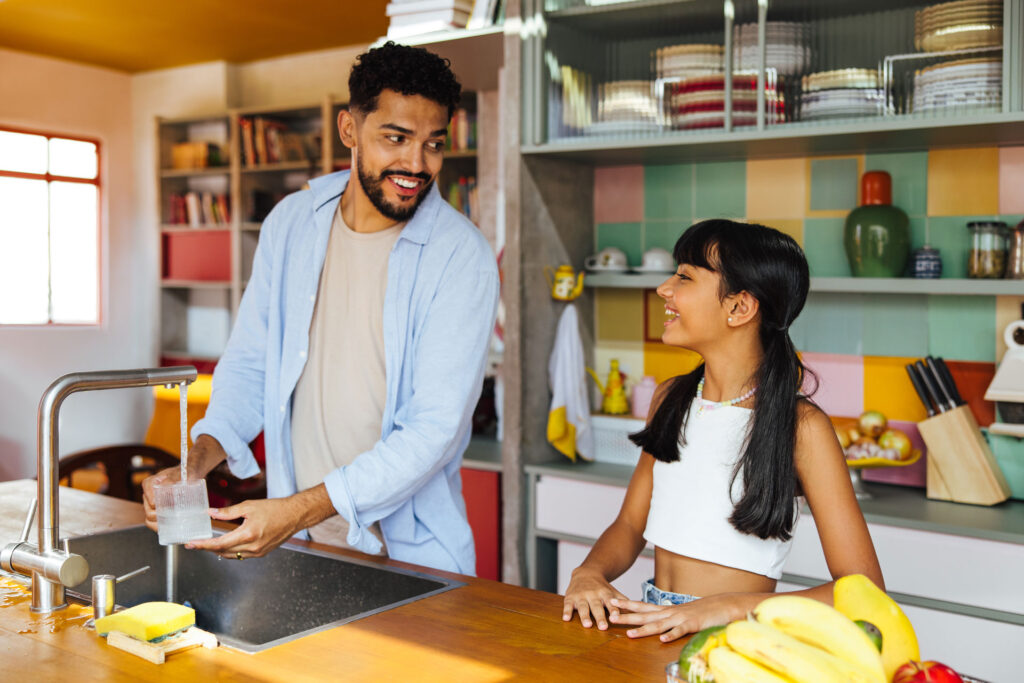 Dark skin bearded father filling water glass in kitchen smiling at dark haired daughter next to him with pigtails.