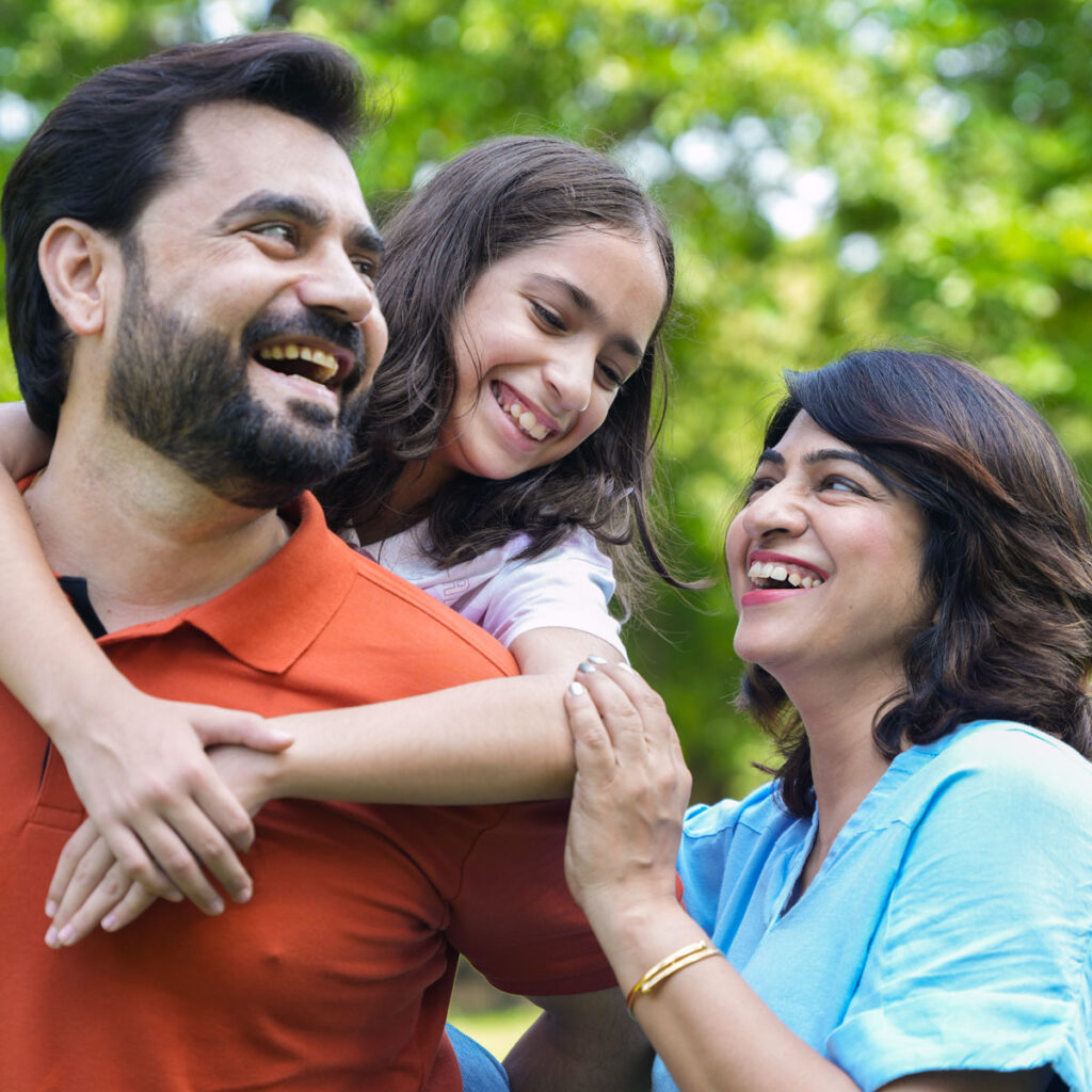 Dark bearded father with daughter on his back and mom standing by their side all smiling in a park.