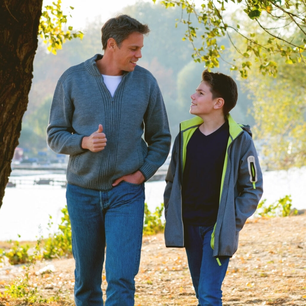 Father and teenage son walking on path near lake and trees looking at each other