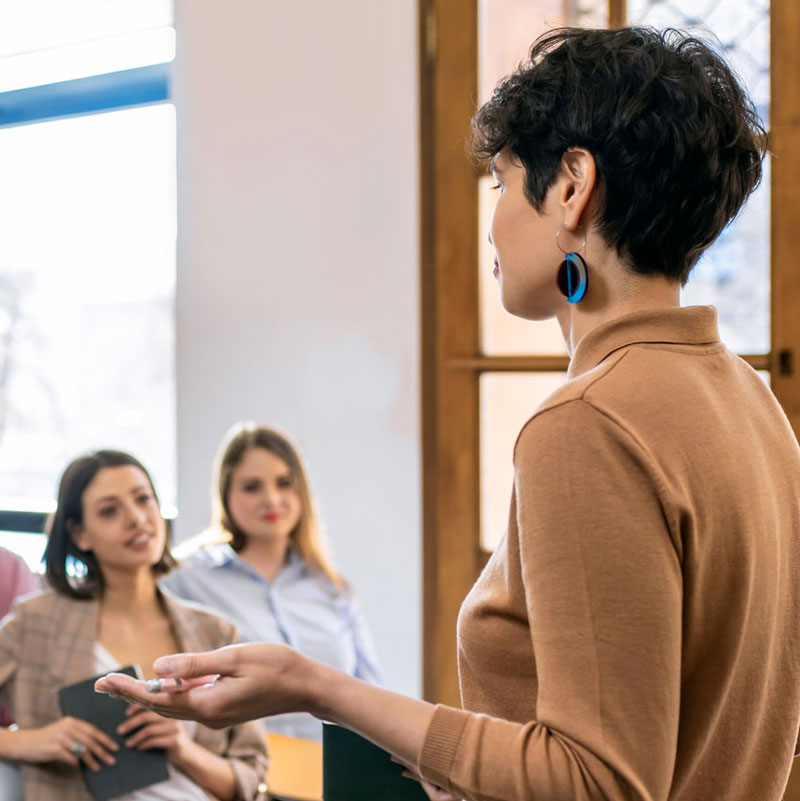 Female lecturer standing and gesturing in front of three women and a man sitting in chairs.