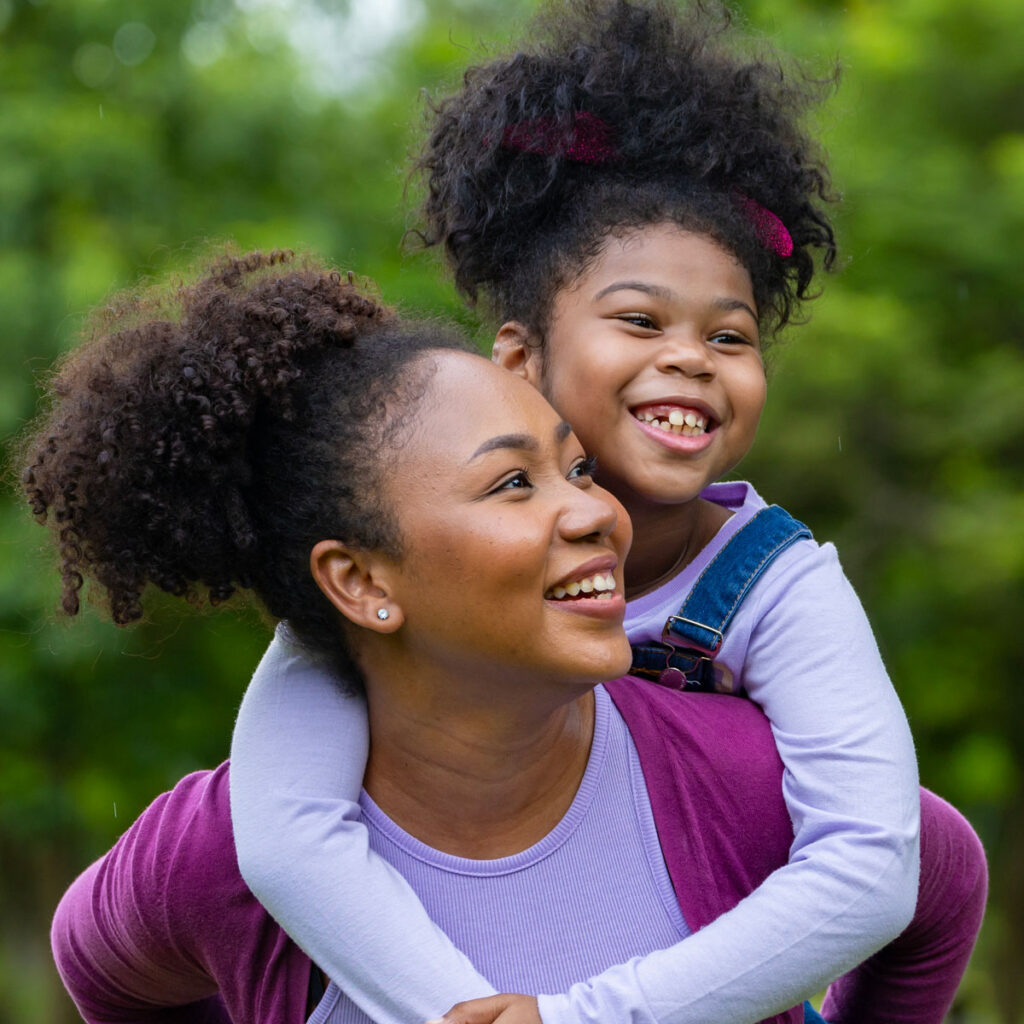 Black mom with daughter on back smiling in a park