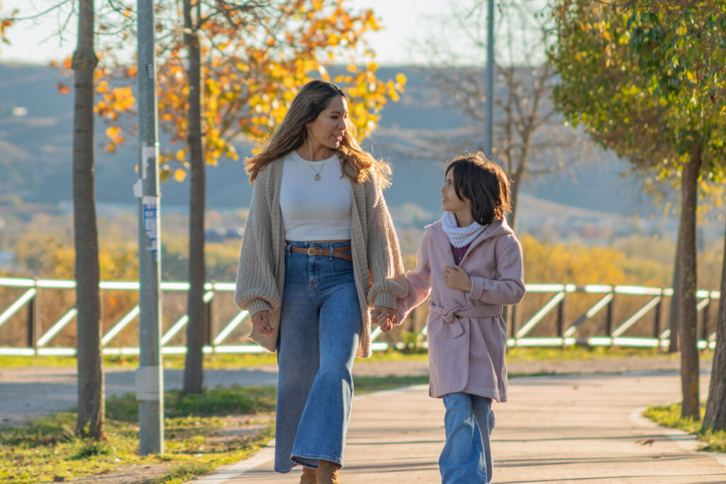 Mom and daughter holding hands and looking at each other while walking down a path with fall trees.