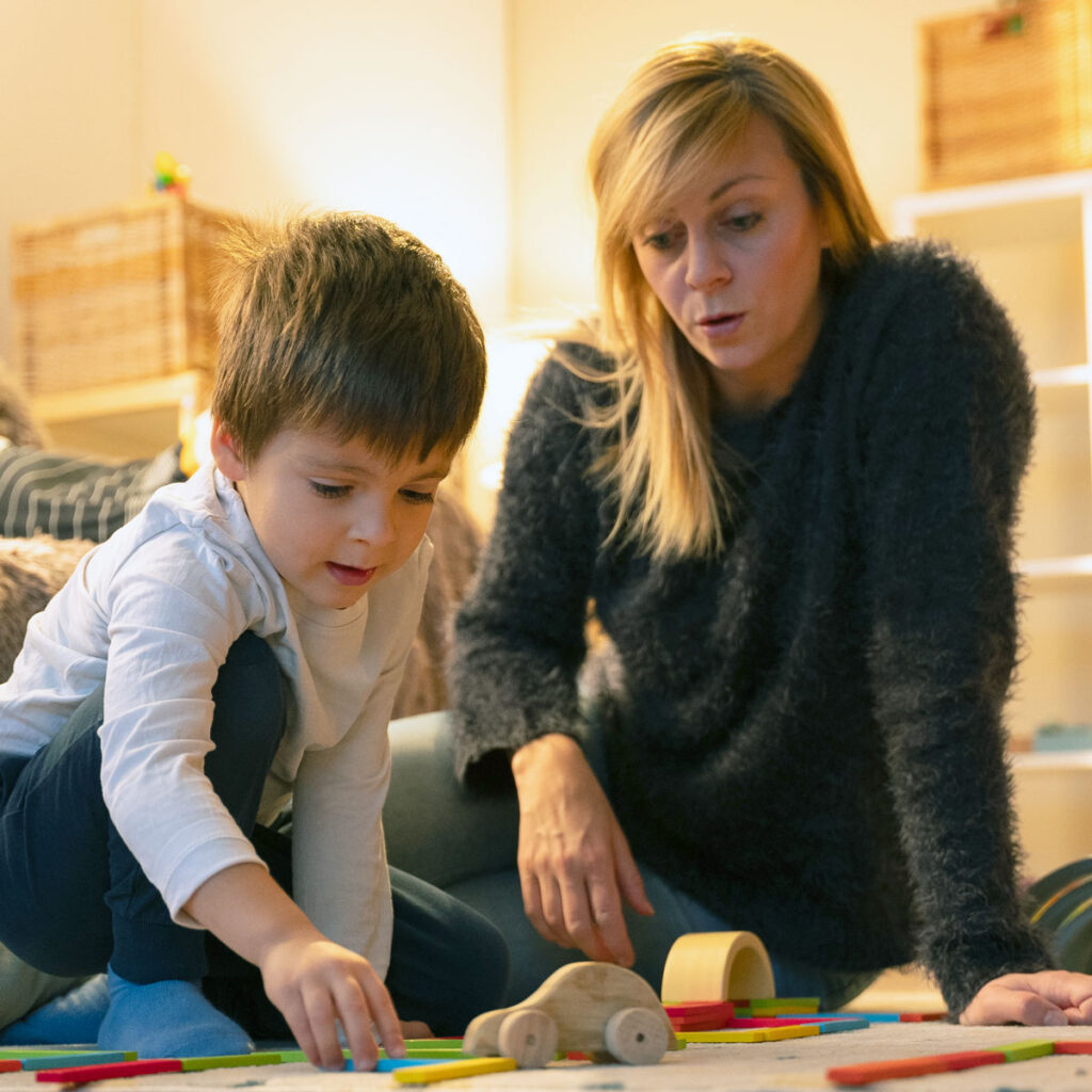 White mom with blonde hair sitting on carpet with young son who is plaing with wood cars and blocks.