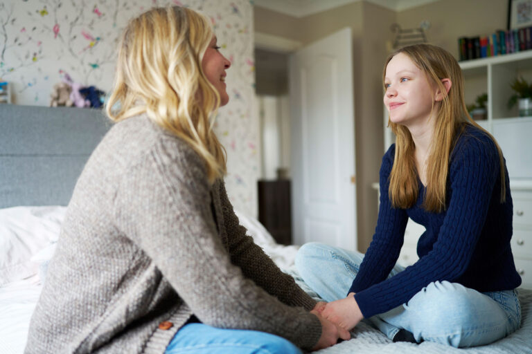 White mother and teenage daughter sitting on bed holding hands looking at each other