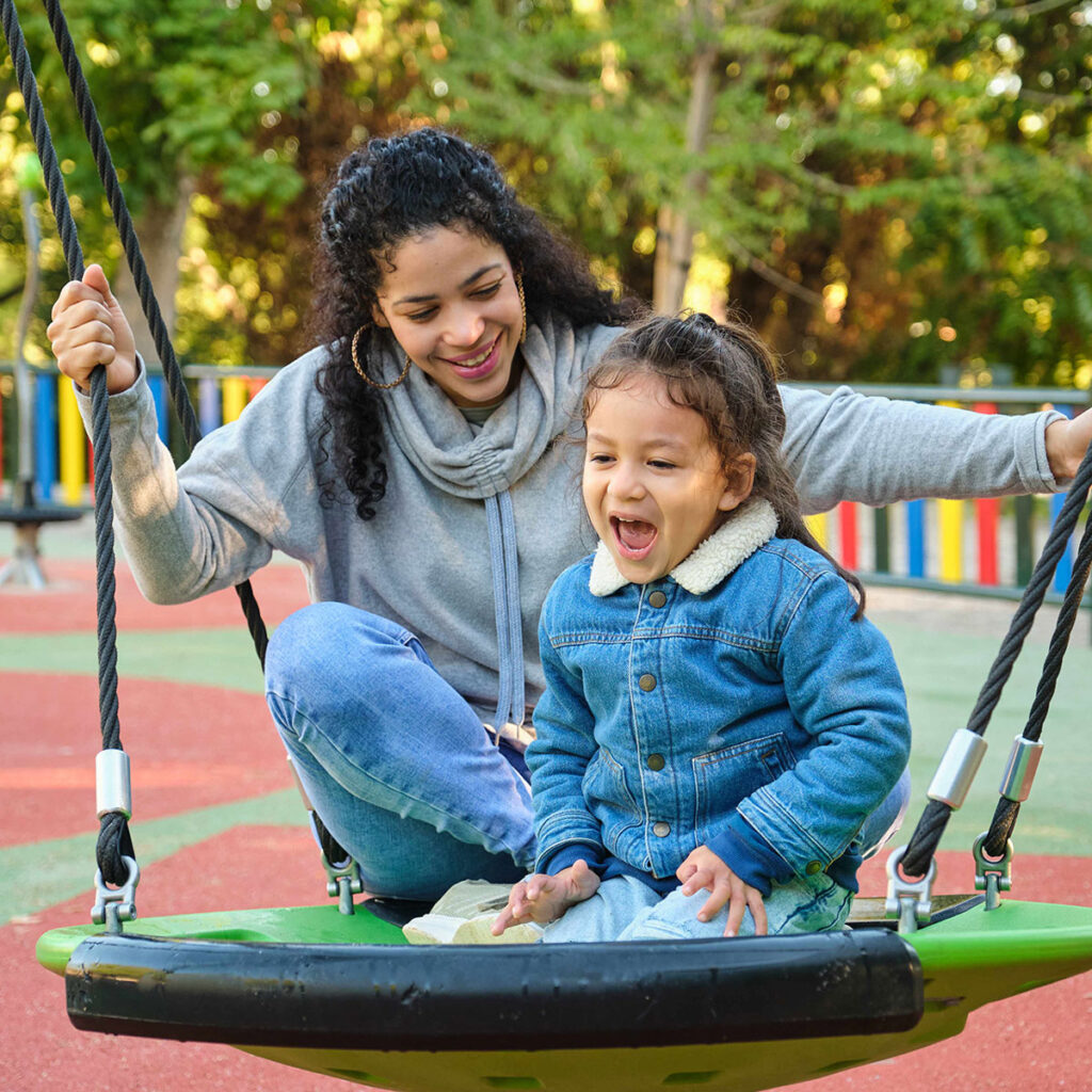 Mother and daughter with dark hair and brown skin sit on a round swing at the playground.